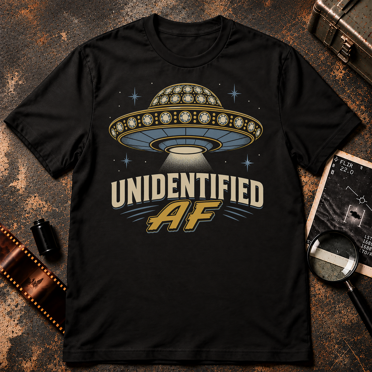 UNIDENTIFIED AF shirt design featuring a jeweled flying saucer decorated with gold and oversized gems, hovering among stars above bold cream and gold lettering on a black background.