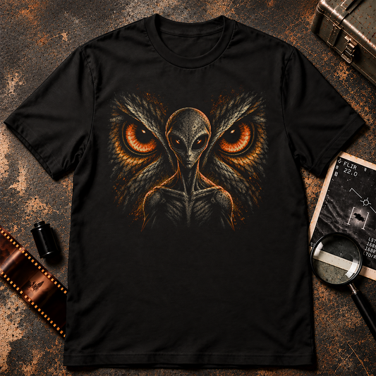 Alien Beneath the Owl's Gaze graphic t-shirt design featuring a sinister grey alien with glowing orange eyes standing before two enormous owl eyes surrounded by detailed feathers on a black background.