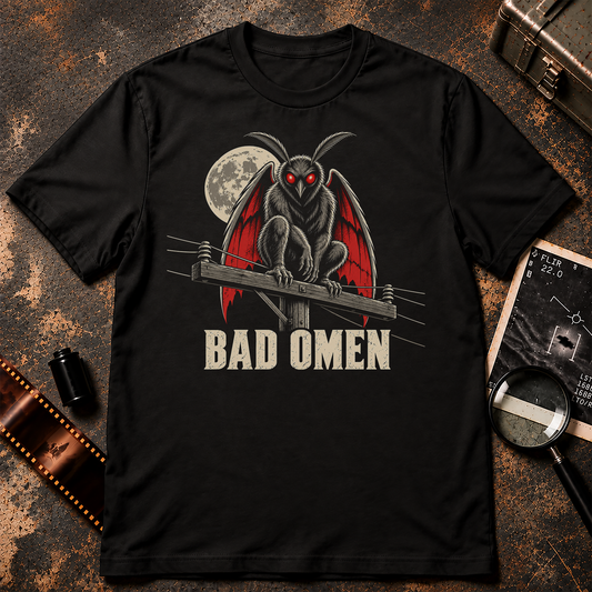 Mothman perched on a wooden power line with glowing red eyes and red wings beneath a full moon above distressed cream text reading Bad Omen.