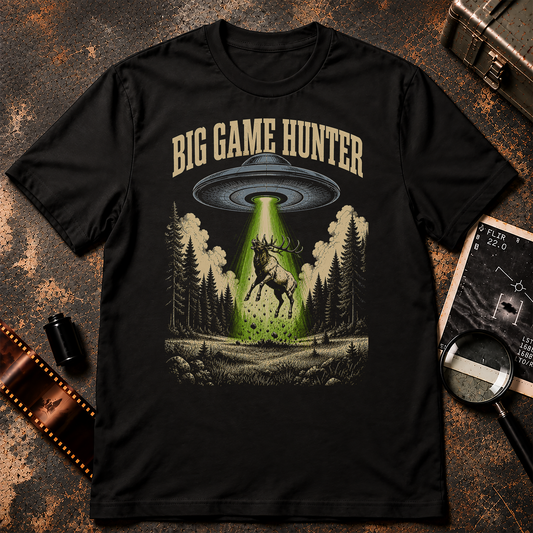 A black short-sleeve graphic t-shirt featuring a 'Big Game Hunter' theme. The graphic shows a green beam from a UFO shining down onto a deer in a forest, with the text 'BIG GAME HUNTER' printed above in a tan serif font.