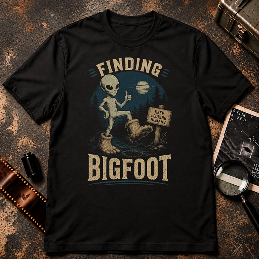Finding Bigfoot shirt design featuring a grey alien wearing oversized Sasquatch feet in a nighttime forest beside a sign reading “Keep Looking Humans.”
