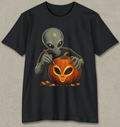 A black short-sleeve crewneck t-shirt featuring a graphic print of a gray alien carving a glowing orange jack-o'-lantern pumpkin, displayed against a textured gray background.