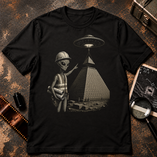 A black short-sleeve crewneck t-shirt featuring a graphic print of a gray alien wearing a construction helmet and safety vest next to a pyramid with a UFO overhead, displayed on a textured gray background.