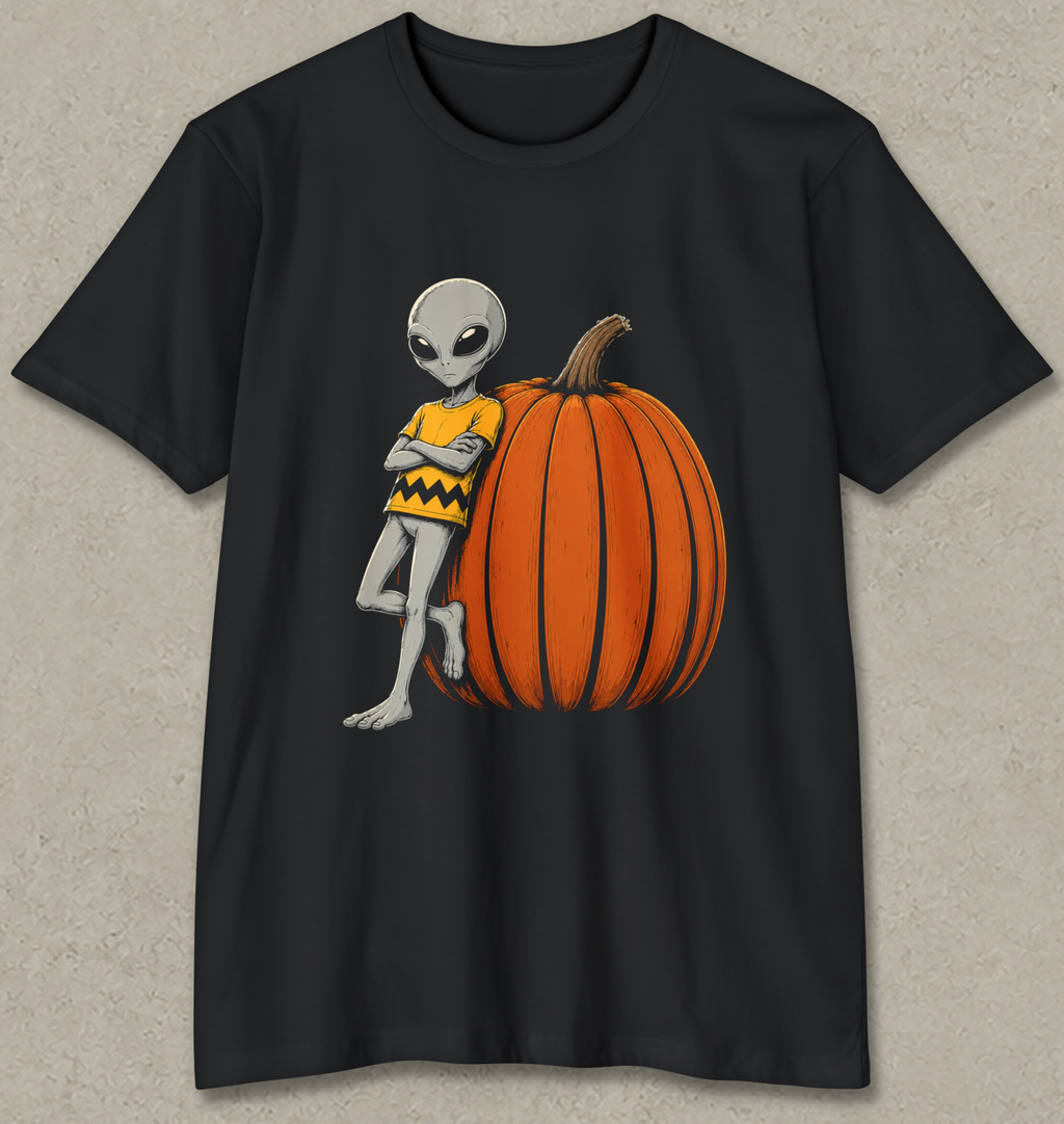 A black short-sleeve crewneck t-shirt featuring a graphic of a gray alien wearing a yellow and black chevron sweater, leaning against a large orange pumpkin.