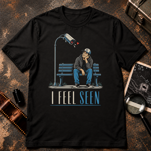 I FEEL SEEN shirt design featuring a man sitting on a park bench gazing affectionately at a surveillance camera leaning toward him with a red heart.