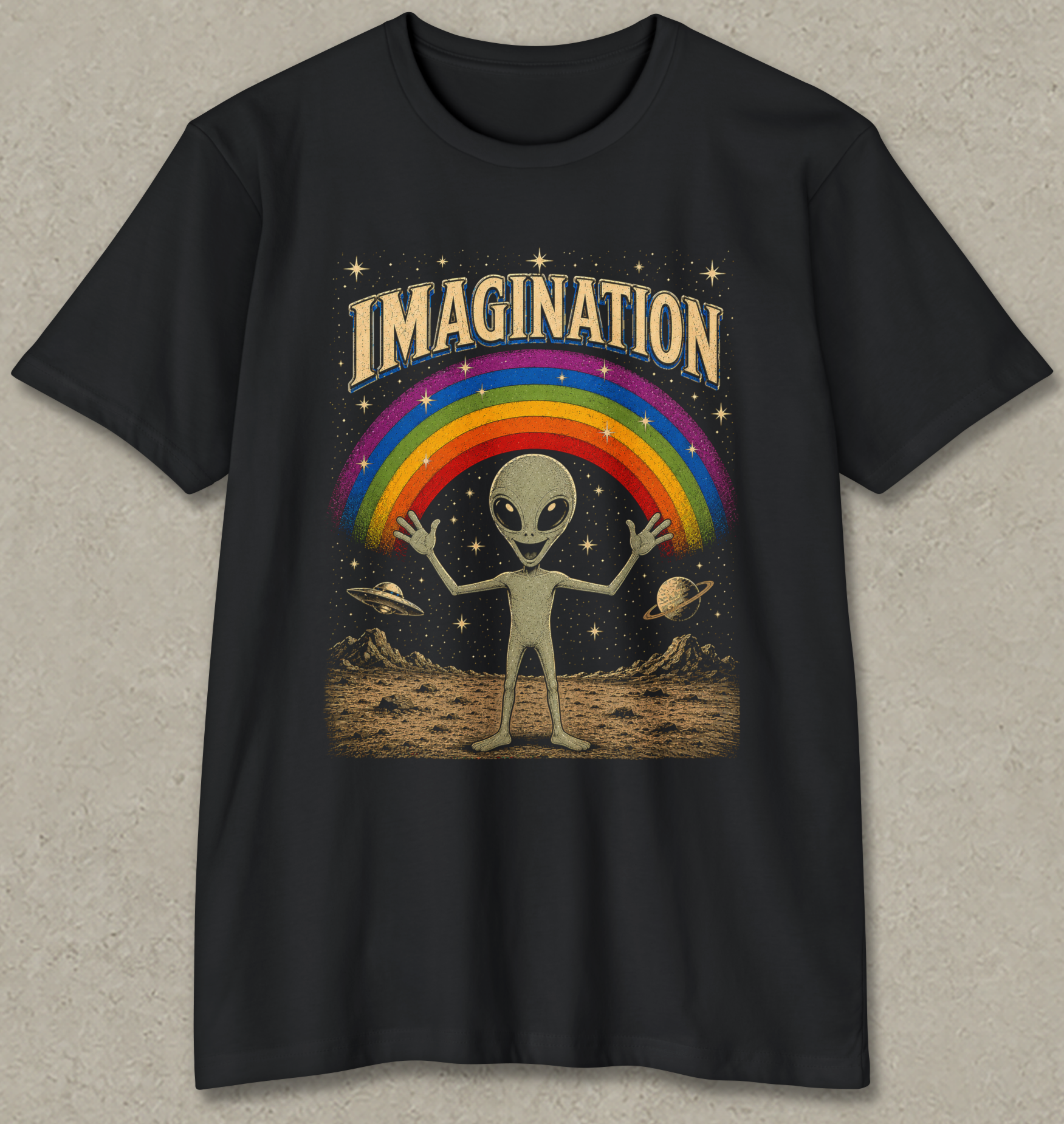 A black short-sleeve t-shirt with a retro-style graphic on the front. The graphic features the word 'IMAGINATION' in a gold-colored arched font above a classic rainbow, a light green alien standing on a dusty surface, and small planets in a starry black sky.