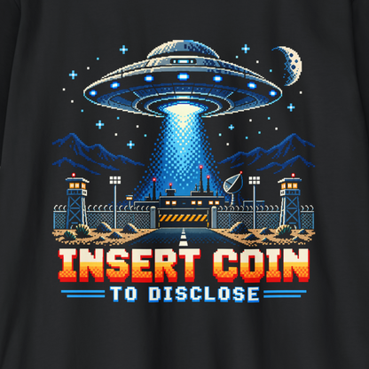 A close-up of a black t-shirt featuring a retro 8-bit pixel art graphic of a UFO landing site with a beam of light, mountains, a moon, and the text 'INSERT COIN TO DISCLOSE' in red and blue.