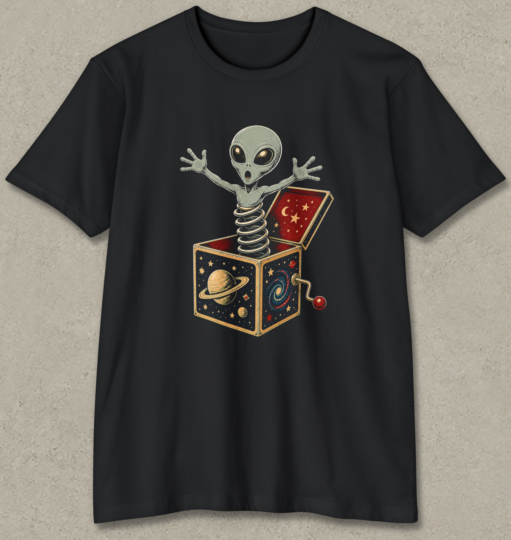 A black short-sleeve crewneck t-shirt featuring a graphic of a grey alien popping out of a juggling box filled with space-themed illustrations, displayed on a textured light gray background.