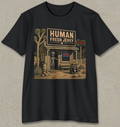 A black short-sleeve crewneck t-shirt with a vintage-style graphic of a desert storefront reading 'HUMAN FRESH JERKY' and featuring a gray alien standing outside, shown against a textured light gray background.