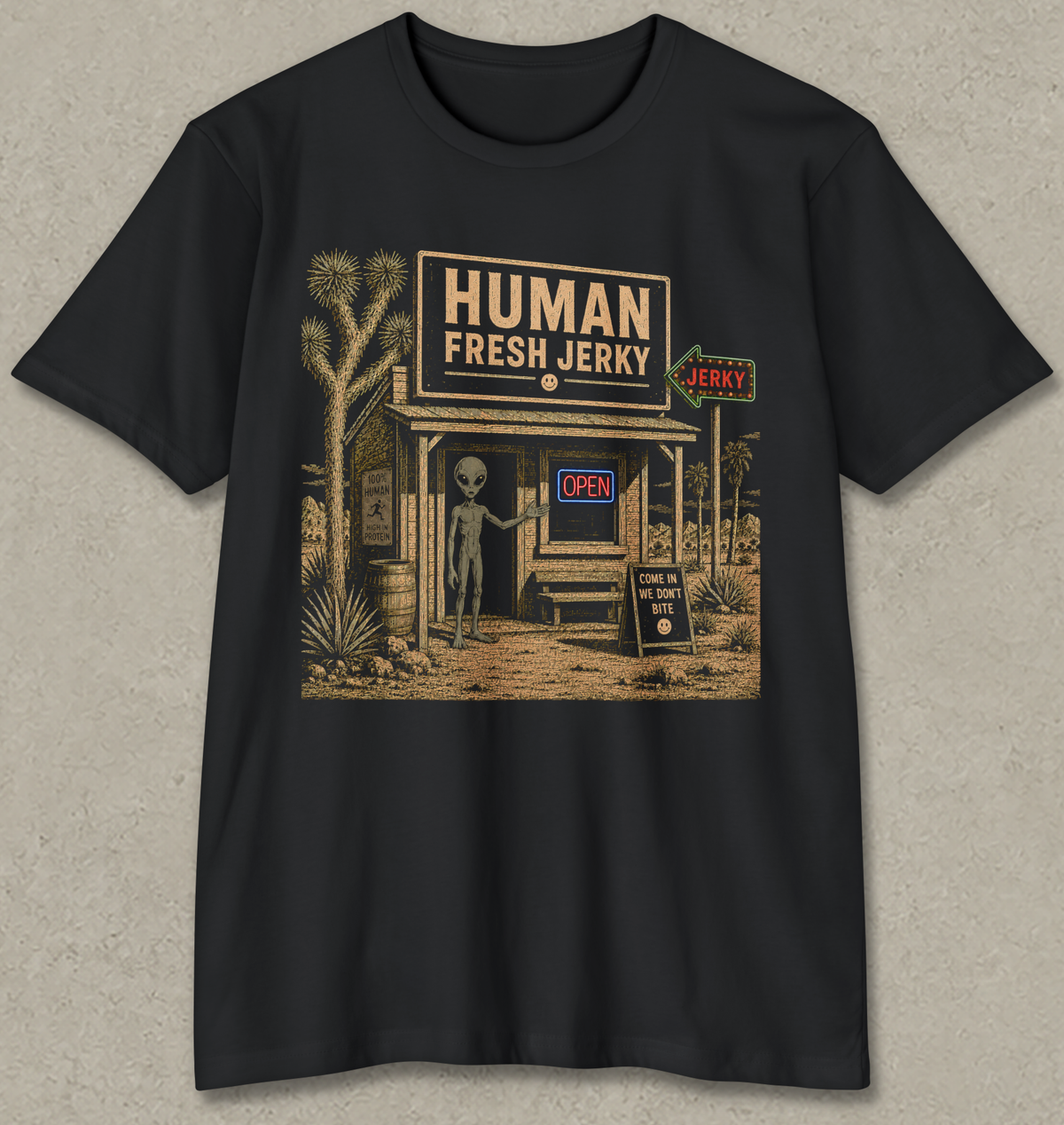 A black short-sleeve crewneck t-shirt with a vintage-style graphic of a desert storefront reading 'HUMAN FRESH JERKY' and featuring a gray alien standing outside, shown against a textured light gray background.