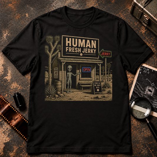 A black short-sleeve crewneck t-shirt with a vintage-style graphic of a desert storefront reading 'HUMAN FRESH JERKY' and featuring a gray alien standing outside, shown against a textured light gray background.