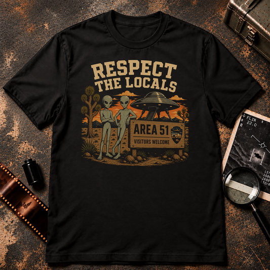 A black short-sleeve crewneck t-shirt with a retro-style graphic on the front. The graphic features two green aliens standing next to a wooden sign that reads 'AREA 51 VISITORS WELCOME' under a sign reading 'RESPECT THE LOCALS'.