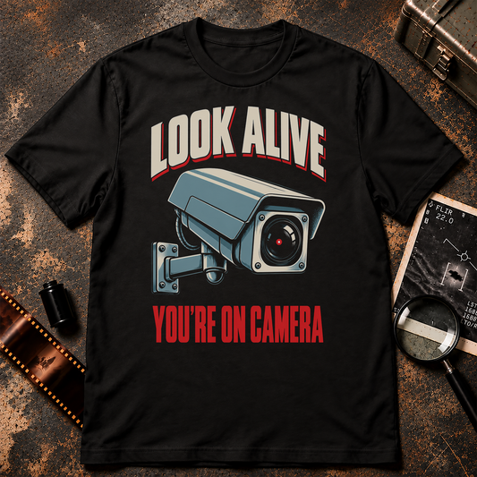 LOOK ALIVE shirt design featuring a large blue-grey surveillance camera with cream highlights and red text reading YOU'RE ON CAMERA.