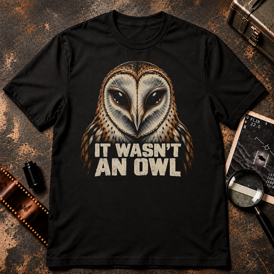 It Wasn't an Owl graphic t-shirt featuring a realistic brown and cream barn owl with oversized glossy black alien eyes. Bold distressed cream lettering reads IT WASN'T AN OWL against a solid black background.