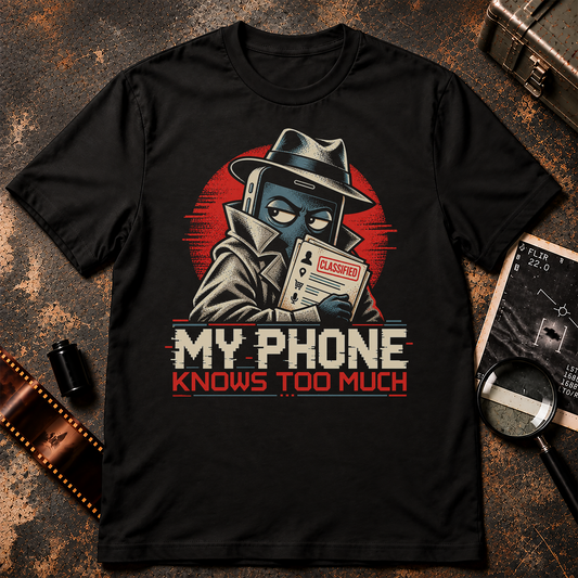 My Phone Knows Too Much graphic t-shirt design featuring a cartoon smartphone disguised as a secret agent wearing a fedora and trench coat while holding a classified dossier. Retro comic illustration in cream, blue, and red with bold MY PHONE KNOWS TOO MUCH lettering on a black background.
