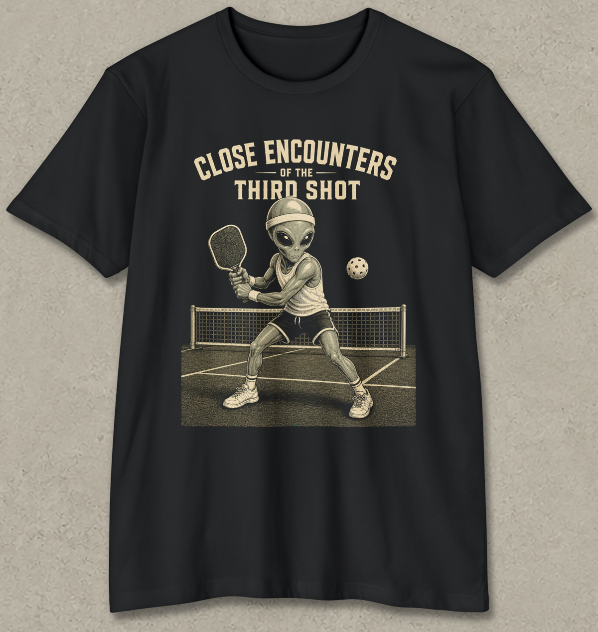 A black short-sleeve crewneck t-shirt with a vintage-style graphic of an alien playing pickleball on a court, with the text 'CLOSE ENCOUNTERS OF THE THIRD SHOT' printed above it. The t-shirt is displayed flat against a textured gray background.