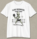 A white short-sleeve crewneck t-shirt with a graphic of a green alien playing pickleball on a court, with the text 'CLOSE ENCOUNTERS OF THE THIRD SHOT' above it. The t-shirt is displayed flat against a textured gray background.