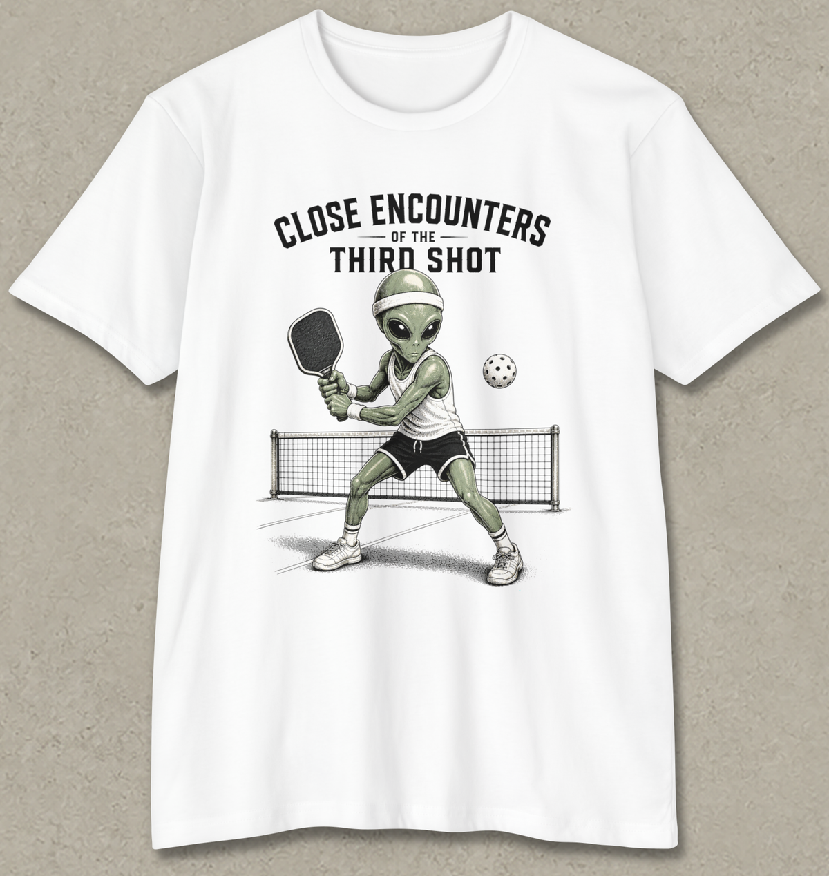 A white short-sleeve crewneck t-shirt with a graphic of a green alien playing pickleball on a court, with the text 'CLOSE ENCOUNTERS OF THE THIRD SHOT' above it. The t-shirt is displayed flat against a textured gray background.