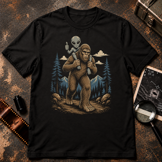 Piggyback shirt design featuring Bigfoot hiking through a mountain forest while carrying a smiling grey alien on his shoulders giving a thumbs-up.