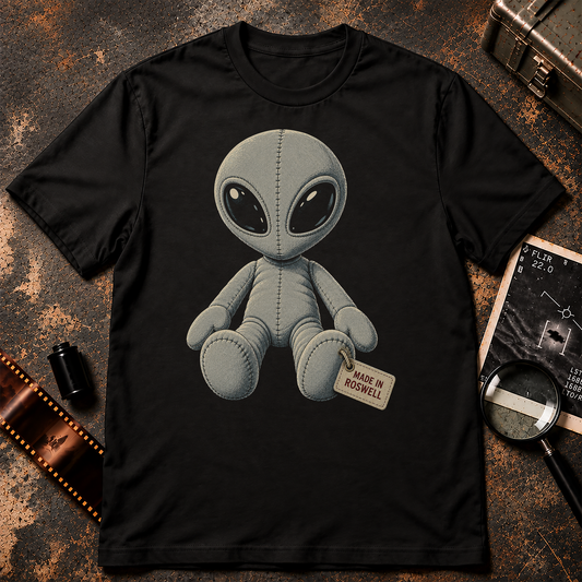 Grey alien plush toy with stitched fabric seams, large black eyes, and a foot tag reading MADE IN ROSWELL on a black background.