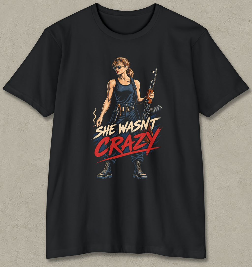 A black short-sleeve crewneck t-shirt with a graphic print of a woman holding an assault rifle, with the text 'SHE WASN'T CRAZY' in stylized red and white lettering.