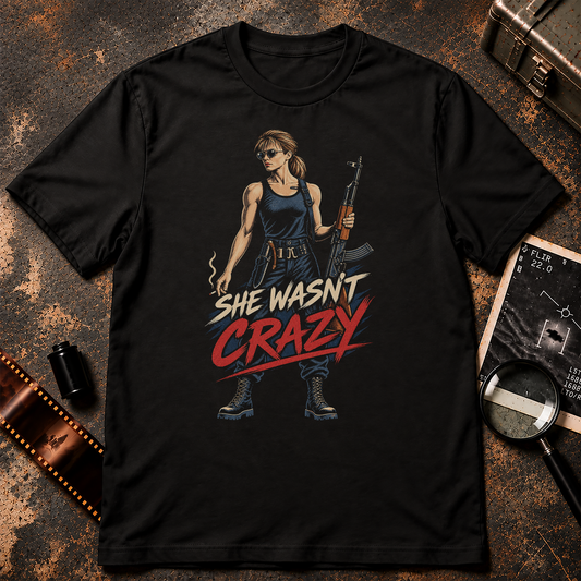 SHE WASN’T CRAZY shirt design featuring a woman in sunglasses and dark tactical clothing holding a rifle and cigarette, with bold cream and red distressed lettering on a black background.