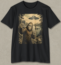 A black short-sleeve crewneck t-shirt with a large vintage-style graphic print on the front. The graphic depicts a Bigfoot taking a selfie in a forest by a lake, with a deer head island in the distance and a flying saucer overhead.