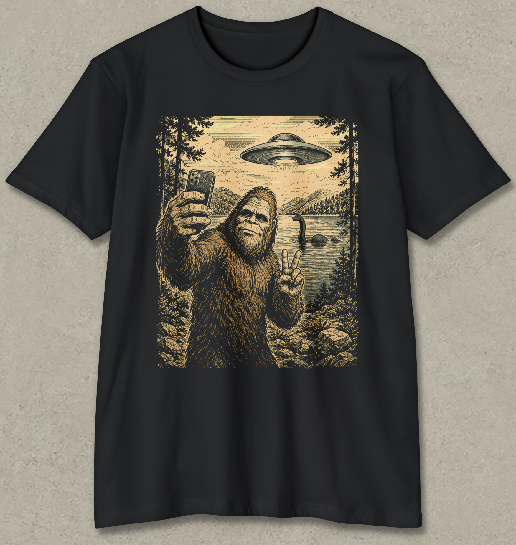 A black short-sleeve crewneck t-shirt with a large vintage-style graphic print on the front. The graphic depicts a Bigfoot taking a selfie in a forest by a lake, with a deer head island in the distance and a flying saucer overhead.
