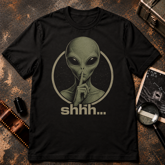 A black short-sleeve crewneck t-shirt featuring a vintage-style graphic of an alien with its finger to its lips inside a circle, with the word 'shhh.