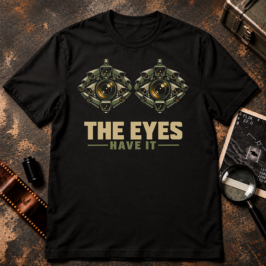 THE EYES HAVE IT shirt design featuring two giant eyes formed from clusters of olive surveillance cameras with glowing amber lenses on black.