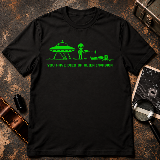 A black short-sleeve crewneck t-shirt with a bright green graphic of an alien, a UFO, and the text 'YOU HAVE DIED OF ALIEN INVASION' printed on the chest, displayed against a textured gray background.