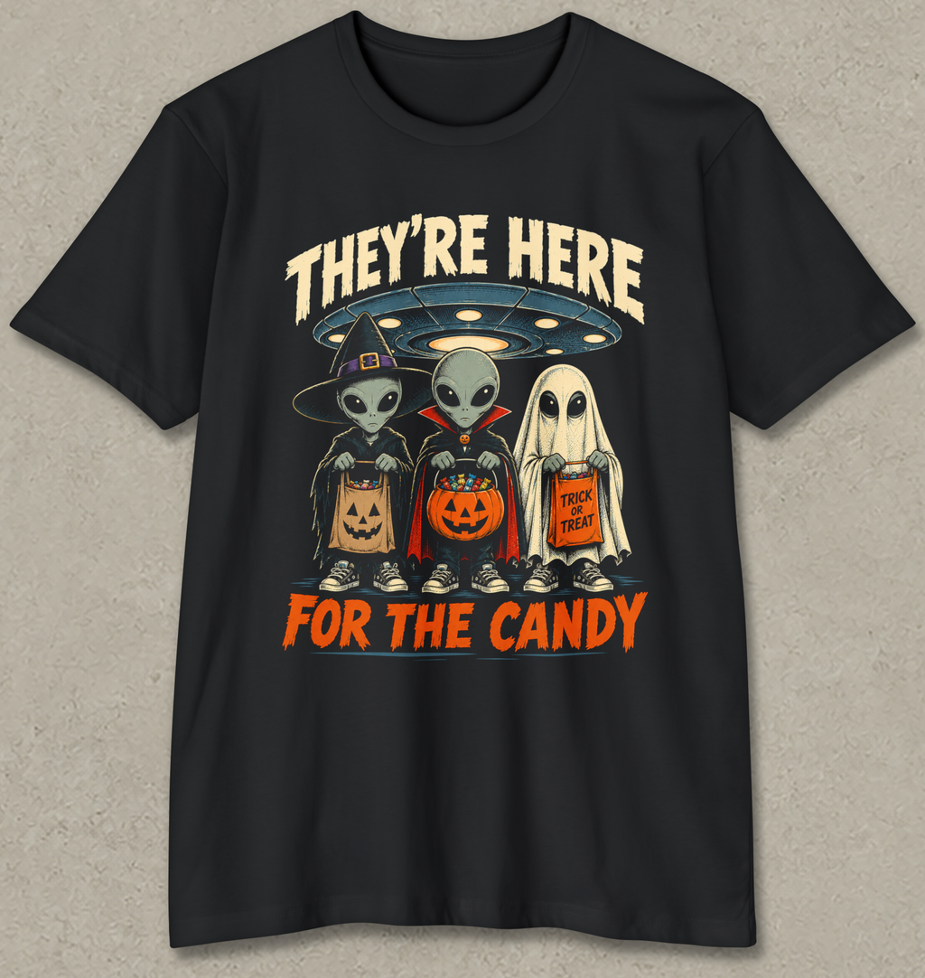 A black short-sleeve crewneck t-shirt with a graphic print of three cartoon aliens dressed in Halloween costumes (wizard, vampire, and ghost) holding jack-o'-lanterns and a candy corn. The text 'THEY'RE HERE' is written in beige above the characters, and 'FOR THE CANDY' is written in orange below.