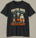 A black short-sleeve crewneck t-shirt with a graphic print of three cartoon aliens dressed in Halloween costumes (wizard, vampire, and ghost) holding jack-o'-lanterns and a candy corn. The text 'THEY'RE HERE' is written in beige above the characters, and 'FOR THE CANDY' is written in orange below.