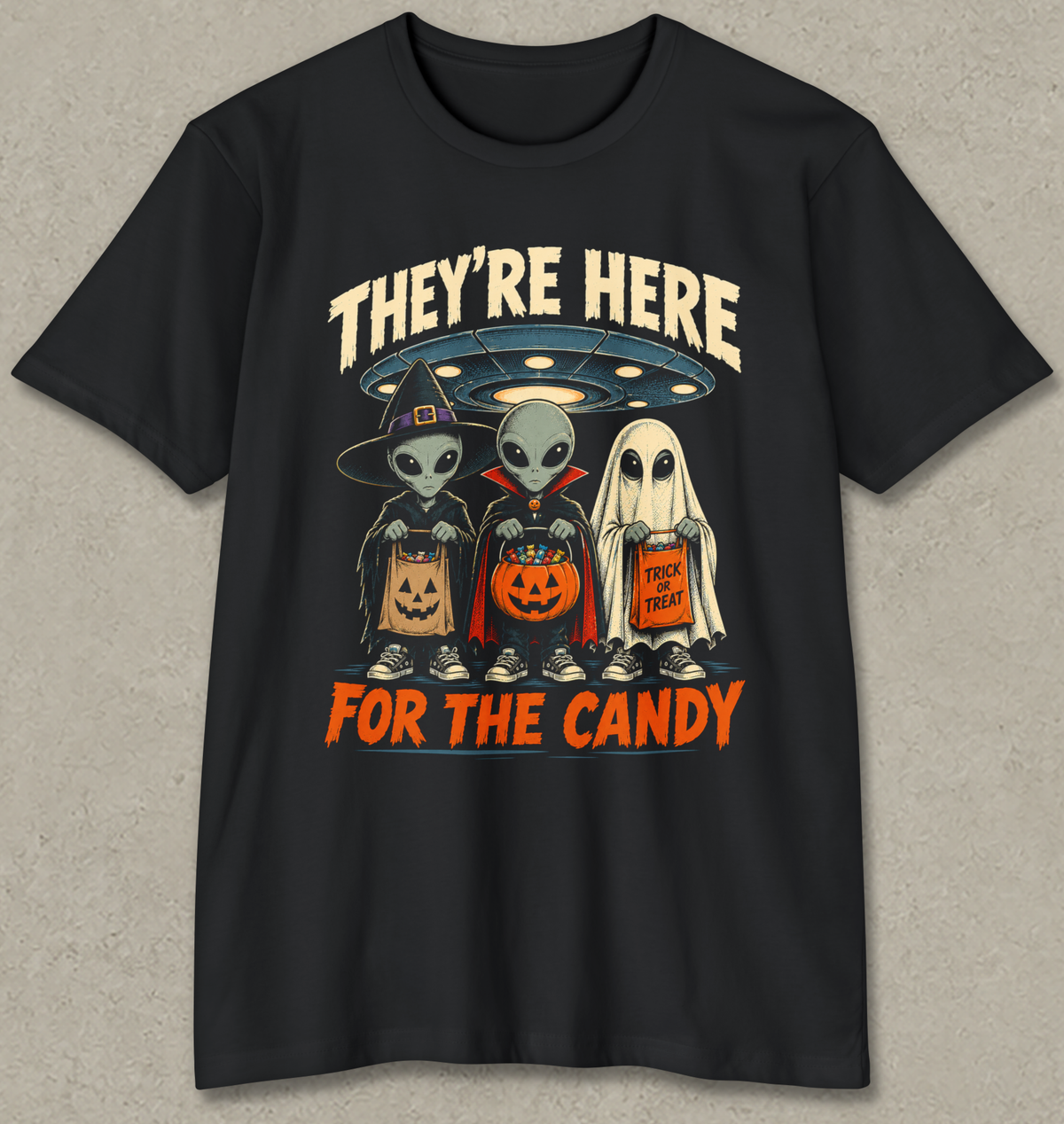 A black short-sleeve crewneck t-shirt with a graphic print of three cartoon aliens dressed in Halloween costumes (wizard, vampire, and ghost) holding jack-o'-lanterns and a candy corn. The text 'THEY'RE HERE' is written in beige above the characters, and 'FOR THE CANDY' is written in orange below.