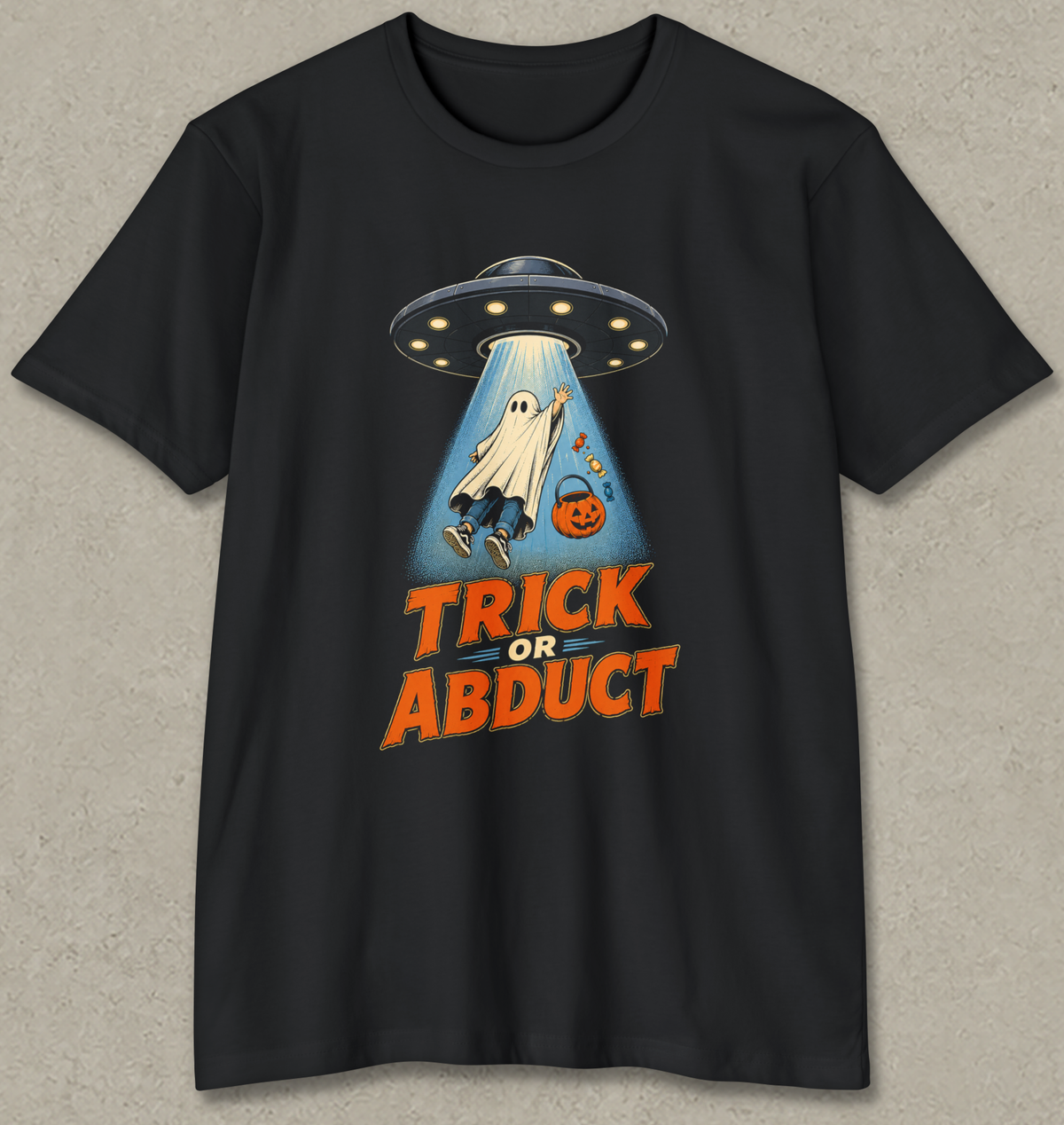 A black short-sleeve crewneck t-shirt with a graphic of a ghost being abducted by a UFO, with the text 'TRICK OR ABDUCT' in orange. The t-shirt is displayed flat against a textured gray background.