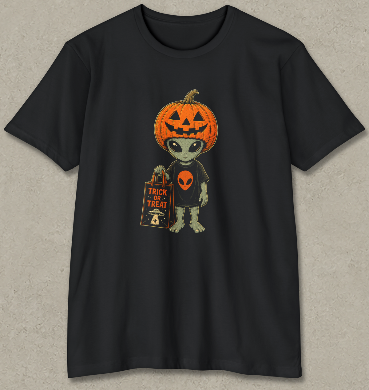 A black short-sleeve crewneck t-shirt featuring a cartoon graphic of a green alien with a jack-o'-lantern head, holding a 'Trick or Treat' bag, displayed on a textured gray background.