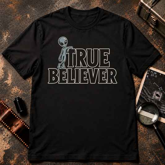 TRUE BELIEVER shirt design featuring a full-body blue-grey alien casually leaning against bold black lettering with cream outlines on a black background.