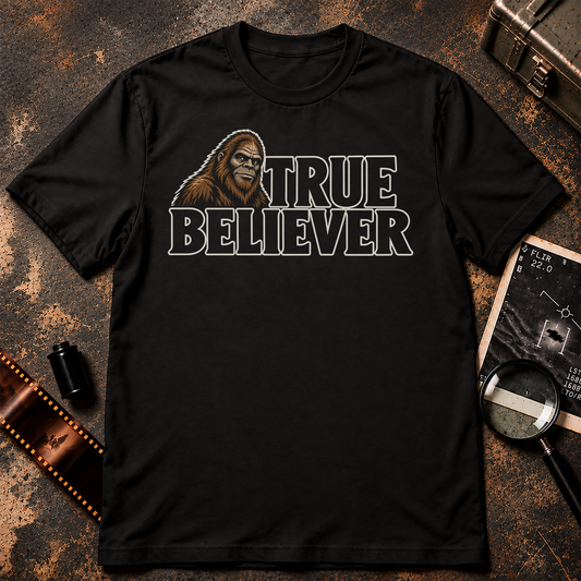 TRUE BELIEVER shirt design featuring a brown Bigfoot portrait beside bold black lettering with cream outlines on a black background.