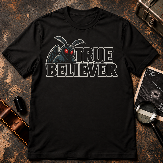 TRUE BELIEVER shirt design featuring a blue-grey Mothman with glowing red eyes beside bold black lettering with cream outlines on a black background.