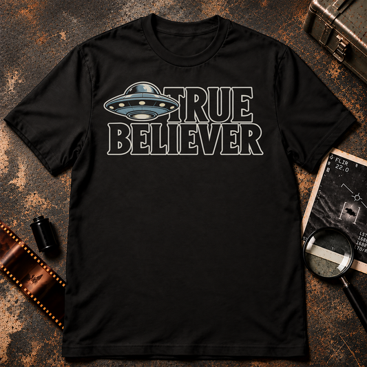 TRUE BELIEVER shirt design featuring a blue-grey flying saucer beside bold black lettering with cream outlines on a black background.