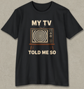 A black short-sleeve crewneck t-shirt with a retro graphic of a television showing a spiral pattern, with the text 'MY TV TOLD ME SO' printed in beige. The t-shirt is displayed flat against a textured gray background.