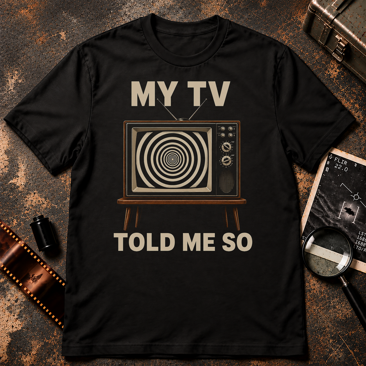 Satirical retro graphic featuring a vintage wood-trim television with antennas and control dials displaying a black-and-cream hypnotic spiral. Large cream text above and below the television reads “MY TV TOLD ME SO” against a black background.