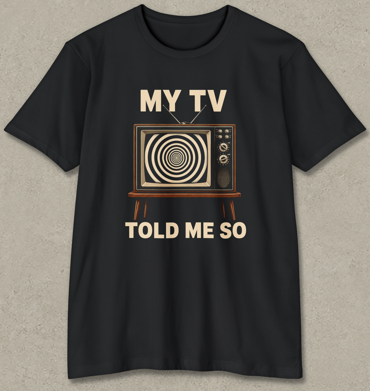 A black short-sleeve crewneck t-shirt with a retro graphic of a television showing a spiral pattern, with the text 'MY TV TOLD ME SO' printed in beige. The t-shirt is displayed flat against a textured gray background.