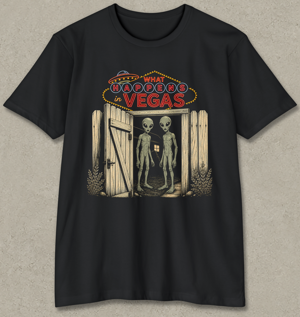 A black short-sleeve crewneck t-shirt with a graphic print on the front. The graphic features a neon sign reading 'WHAT BROOKERS IN VEGAS' above an open wooden door with two tall, thin gray aliens standing inside.