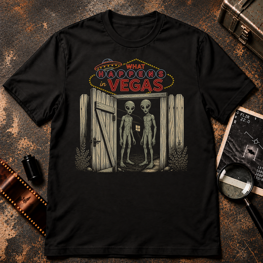 A black short-sleeve crewneck t-shirt with a graphic print on the front. The graphic features a neon sign reading 'WHAT BROOKERS IN VEGAS' above an open wooden door with two tall, thin gray aliens standing inside.