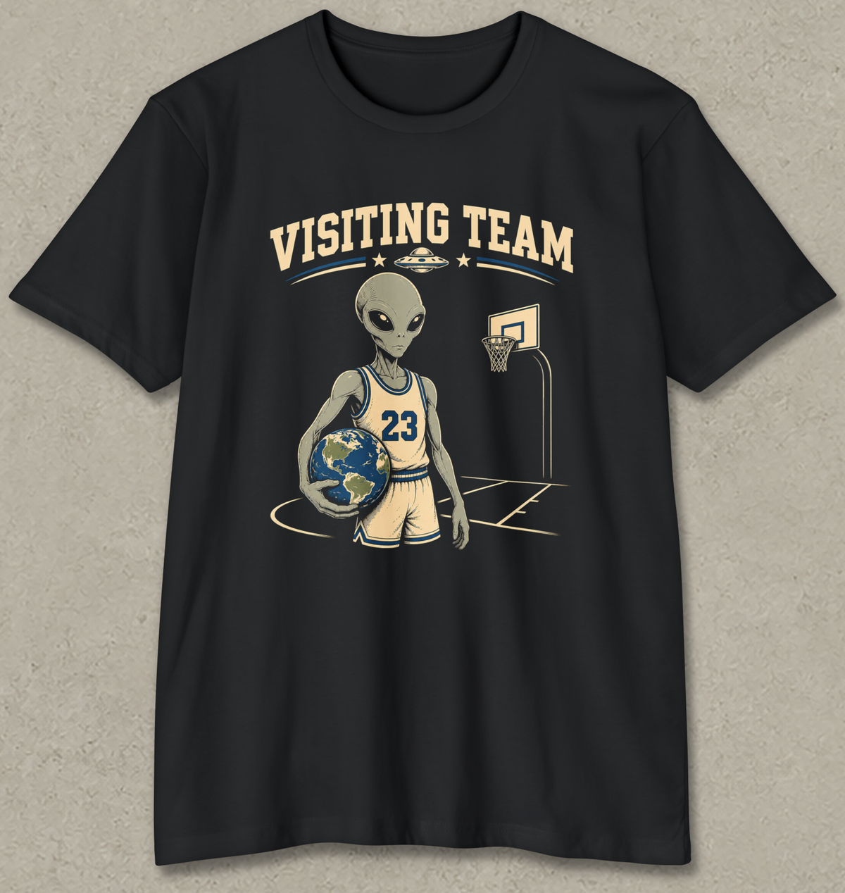 A black short-sleeve crewneck t-shirt featuring a graphic of a cartoon alien basketball player holding a globe, with the text 'VISITING TEAM' above him, displayed against a textured gray background.