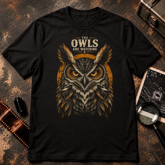 The Owls Are Watching T-Shirt graphic featuring a fierce owl with glowing amber eyes, dramatic cream and orange feathers, and two hidden surveillance cameras emerging from its body. Distressed lettering above reads THE OWLS ARE WATCHING against a black background.