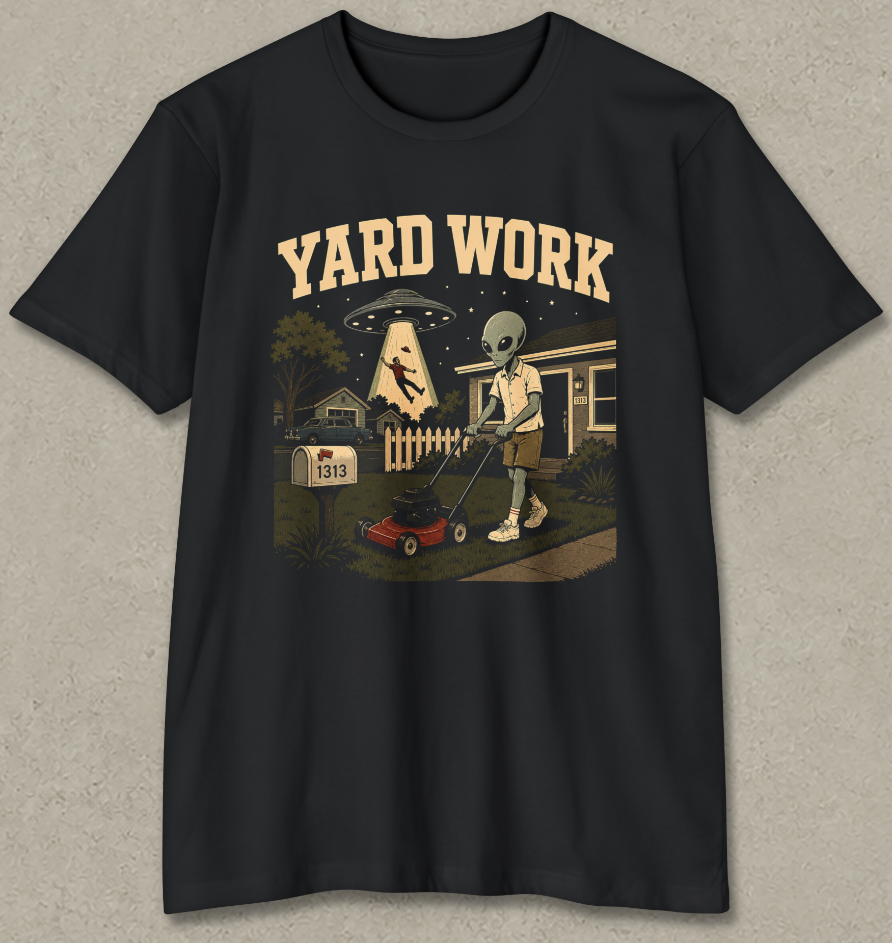 A black short-sleeve crewneck t-shirt with a vintage-style graphic on the front. The graphic features the text 'YARD WORK' in a retro cream font above an illustration of an alien mowing a lawn with a red lawnmower, with a UFO and house in the background.